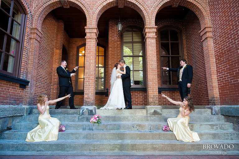 wedding party pointing at bride and groom kissing under archway