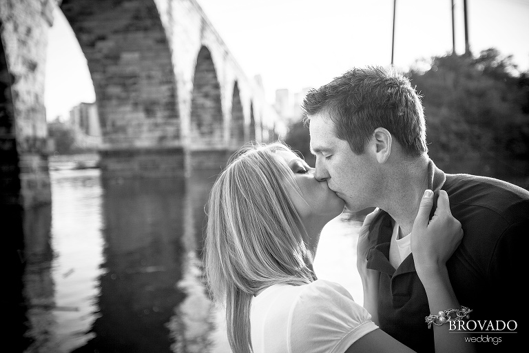 black and white photography of engaged couple kissing in front of stone arch bridge