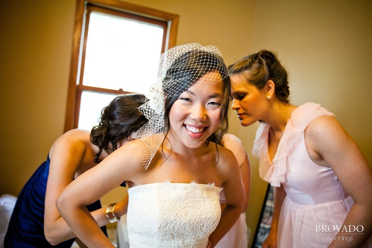 bride smiling through her mesh veil while bridesmaids help put her dress on