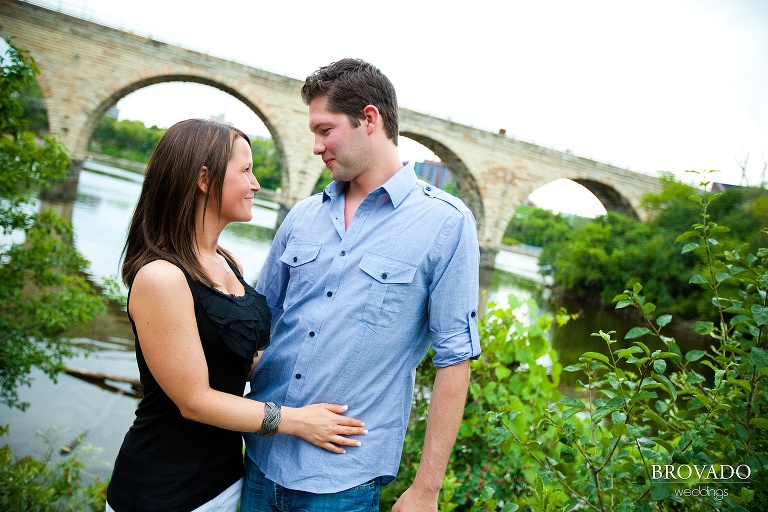 fiances smiling at each other in front of stone arch bridge in downtown minneapolis