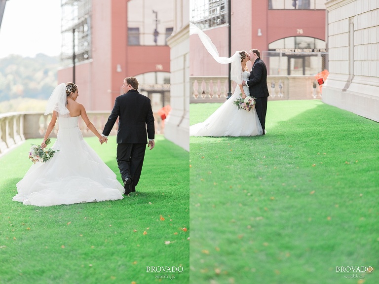 Bride and groom walk across green grass, veil tossed in the wind
