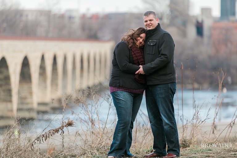 Woman leans on her fiance in front of stone arch bridge
